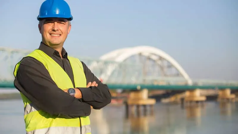 Construction worker smiling in front of bridge in the background.