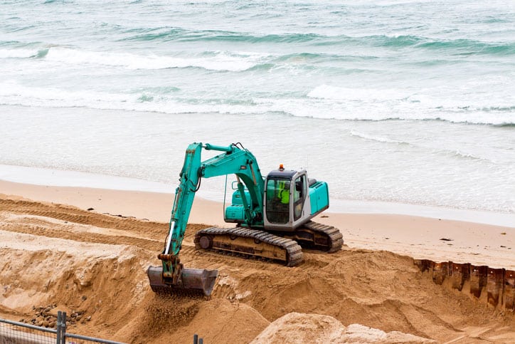 Green excavator digging on the beach with ocean, full frame horizontal composition